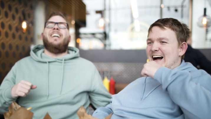 Ung man med assistent skrattar vid restaurangbord med hamburgare