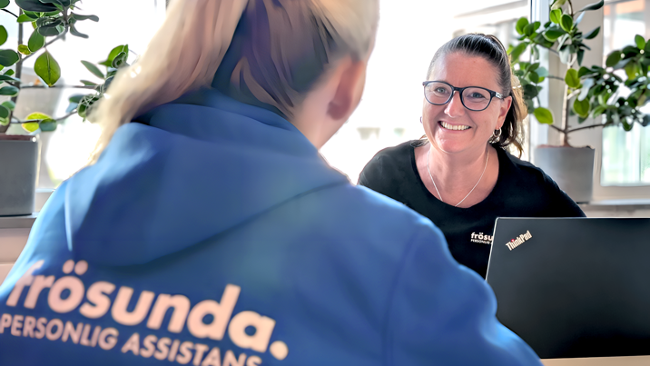 Person i Frösunda-hoodie möter leende medarbetare med glasögon vid laptop på kontor