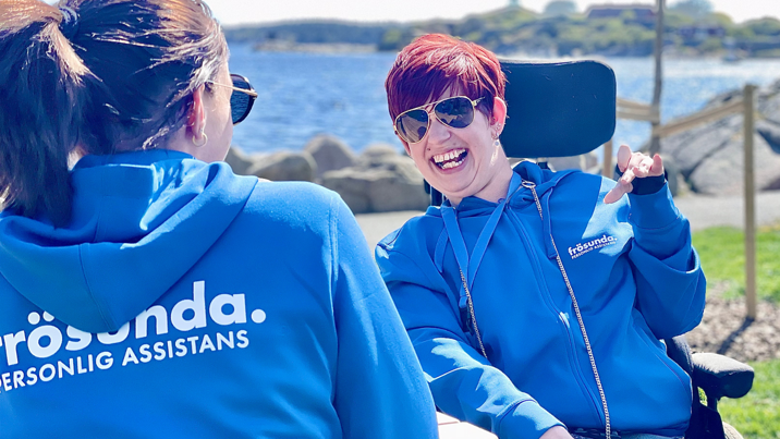 Leende kund i rullstol och assistent i blå Frösunda-hoodies skrattar tillsammans vid vattnet