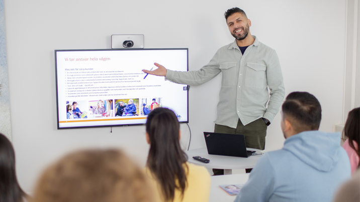 Man håller presentation för grupp kollegor i mötesrum med skärm.
