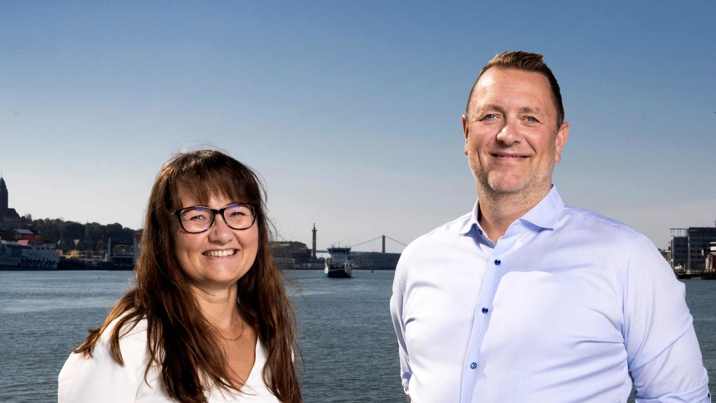 Kvinna med glasögon och man i ljusblå skjorta ler framför hamninloppet med bro i bakgrunden