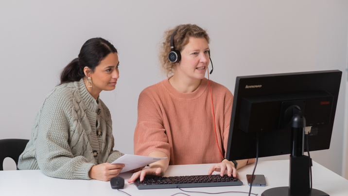 Två kvinnliga kollegor arbetar vid datorskärm, den ena med headset
