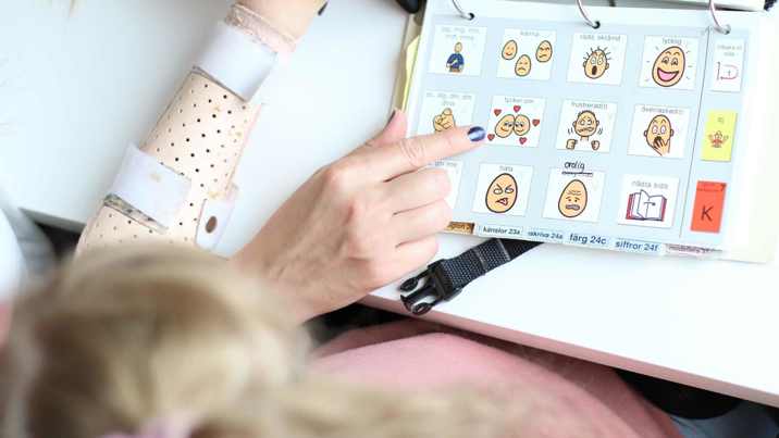 Hand pekar i kommunikationspärm med bildsymboler för känslor medan flicka tittar på