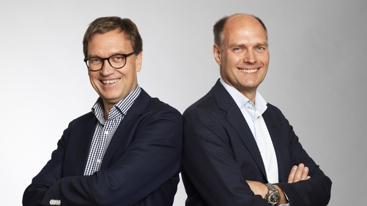 Två leende män i mörka kavajer står rygg mot rygg med armarna i kors i studio