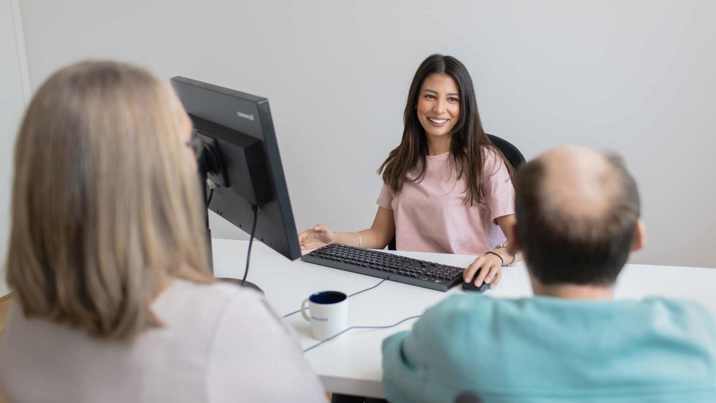 Leende kvinnlig medarbetare tar emot två besökare vid skrivbord på kontor.