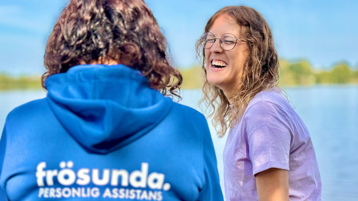 Personlig assistent i Frösunda-hoodie samtalar med leende kund utomhus vid vattnet.